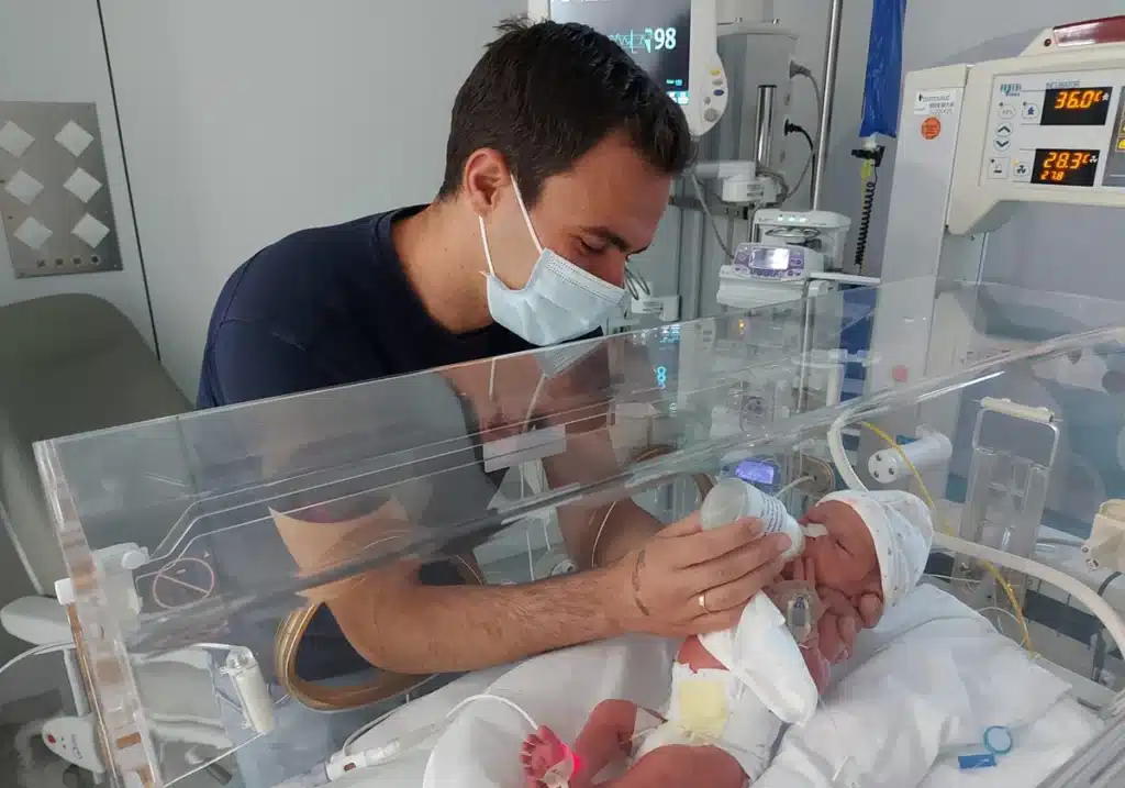 Víctor, con Victoria, en la UCI, poco después del nacimiento de la pequeña. Su papa le esta dando el biberón, Victoria se encuentra dentro de una incubadora